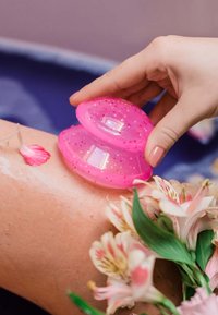 Różowy silikonowy kubek do masażu z brokatem, trzymany nad nogą. W scenie widoczne są świeże kwiaty oraz wilgotna, błyszcząca powierzchnia skóry.