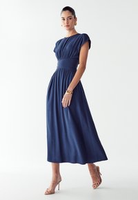 Mornarsko modra maksi obleka s kratkimi rokavi, nabranim zgornjim delom in prilagojenim pasom. Nosimo jo z bež sandali na visokem podplatu.