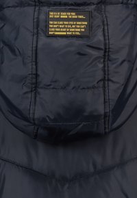 Zateplená bunda v námořnické modři s kapucí, diagonálním prošíváním a černou nálepkou se žlutým textem na zadní straně.