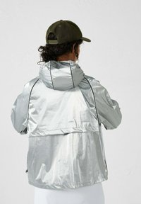 Zilveren waterdichte jas met capuchon, met een losse pasvorm, zichtbare naden en een lichtgewicht, glanzende textuur. Draagt een groene pet.