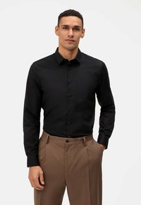 Camicia nera a maniche lunghe con design a bottoni, caratterizzata da un colletto e una texture liscia, abbinata a pantaloni marrone chiaro.