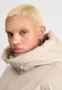 Doudoune beige avec col montant, fermeture éclair et texture lisse, portée avec des boucles d'oreilles créoles. Gros plan sur les traits du visage.