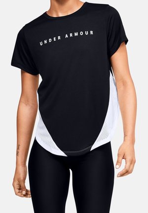 Person trägt ein schwarzes und weißes kurzärmeliges Under Armour Sportshirt und schwarze Leggings, steht vor einem einfarbigen Hintergrund.