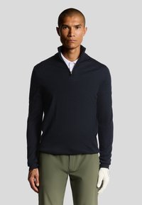Pullover blu scuro con zip a quarti, tessuto testurizzato, maniche lunghe, indossato sopra una camicia con colletto bianca, abbinato a pantaloni verde chiaro e un guanto da golf.