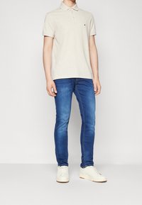 Beige poloko s krátkým rukávem a malým logem, doplněné modrými slim fit džíny a bílými teniskami.