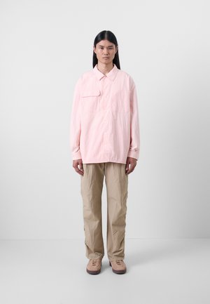 Chemise boutonnière oversize rose pâle avec une poche poitrine, associée à un pantalon texturé beige et des chaussures beiges, se tenant sur un fond uni.