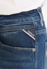 Tummansiniset farkut, joissa on teksturoitu pinta ja selkeä valkoinen logo-osa, jossa lukee "REPLAY", takataskussa.