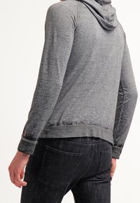 Felpa grigia con finitura testurizzata, con cappuccio regolabile a coulisse e orlo inferiore leggermente sagomato, abbinata a jeans in denim scuro.