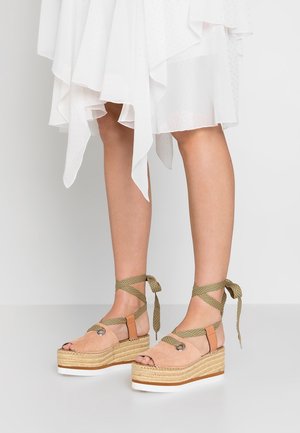Benen met beige sleehak espadrille sandalen met enkelriempjes en een witte asymmetrische zoom jurk.