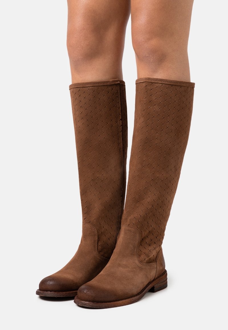 Bottes hautes en daim marron avec des détails texturés portées sur des jambes nues, posées sur un fond blanc.
