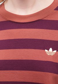 Burgundijas un rūsas svītrota kokvilnas T-kreklu ar ribotu apaļu kakla izgriezumu un nelielu baltu Adidas logotipu krūtīs. Mīksts audums.