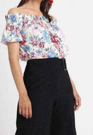Femme portant un chemisier à fleurs décolleté aux épaules avec des fleurs rouges, bleues et roses, assorti à un pantalon noir taille haute avec une boucle latérale.