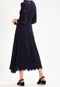 Granatowa sukienka koronkowa z długimi rękawami i falbaniastym dołem, zdobiona kwiatowymi wzorami, o swobodnym kroju, uzupełniona butami na blokowym obcasie.