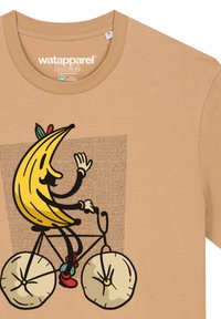 Tan t-shirt met een cartoon banaan karakter dat op een fiets met grote, ronde wielen rijdt. Het ontwerp bevat zwarte omlijning en kleurrijke accenten.