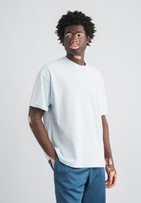 Ανοιχτό μπλε βαμβακερό μπλουζάκι με στρογγυλή λαιμόκοψη, κοντά μανίκια και μικρό λευκό λογότυπο στο στήθος, σε συνδυασμό με μπλε παντελόνια.