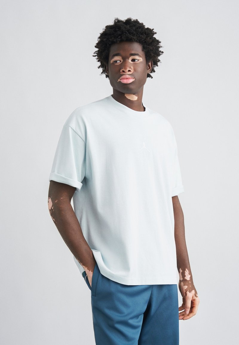 Ανοιχτό μπλε βαμβακερό μπλουζάκι με στρογγυλή λαιμόκοψη, κοντά μανίκια και μικρό λευκό λογότυπο στο στήθος, σε συνδυασμό με μπλε παντελόνια.