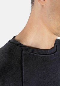 Svart hettegenser med en strukturert overflate og ribbet hals. Nærbilde viser detaljer ved hals og skulder, som fremhever materialet og sømmen.