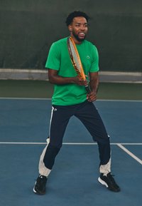 Grønn kortermet skjorte og marineblå joggebukser med hvite side-striper. Svarte tennissko. Holder en tenniskølle, klar til å spille.