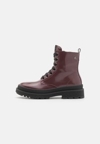 Burgunderfärgade glänsande läderankelboots med svarta runda snörningar, en tjock svart sula och en dragflik. Har en logo på sidan.