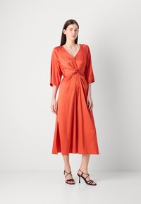 Rochie portocalie din satin cu decolteu în V, talie legată și mâneci trei sferturi, cu o fanta pe lateral. Modelul este asortat cu sandale negre cu curele.