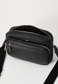 Black pebbled leather crossbody bag with two zippered compartments and adjustable black fabric strap on white surface.
