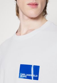 Balts kokvilnas T-krekls ar zilu taisnstūra grafiku, kurā rakstīts "KARL LAGERFELD JEANS" baltā krāsā. Gluda tekstūra un apaļa kakla dizains.