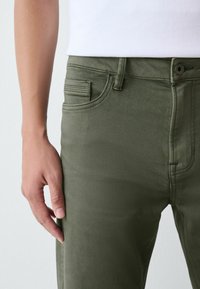 Õlivärvi puuvillased püksid, millel on vormitud disain, viie-tasku stiil ja peened õmblusdetailid. Käsi toetub õrnalt reie piirkonda.
