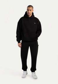 Zwarte hoodie en joggingbroek set gemaakt van zachte stof. De hoodie heeft een zak aan de voorkant en een logo-detail; beide hebben een ontspannen pasvorm. Witte sneakers.