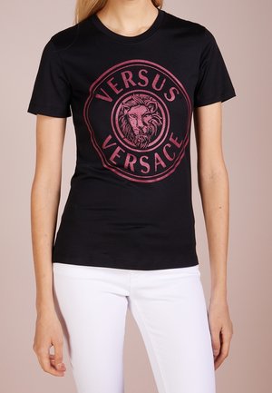 Czarny dopasowany t-shirt z dużym różowym okrągłym logo przedstawiającym głowę lwa oraz napisem "Versus Versace", noszony z białymi spodniami.