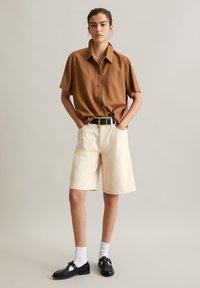 Bruin overhemd met korte mouwen en knoopsluiting, crèmekleurige knielange shorts, zwarte riem en zwarte schoenen met witte sokken. Eenvoudig, schoon ontwerp.