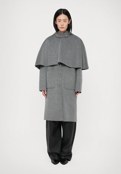 Manteau en laine gris avec un design de cape, décoré de boutons dorés et de poches latérales. Le tissu a une texture douce et est complété par un large pantalon noir.