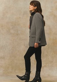 Kvinde i sorte støvler, sorte bukser og en sort-hvid houndstooth blazer står sidelæns mod en beige tekstureret baggrund.