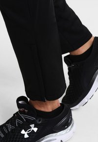 Pantalons de sport noirs avec une coupe ajustée, dotés d'un détail zippé au niveau des poignets, associés à des chaussures de sport noires avec un design texturé.
