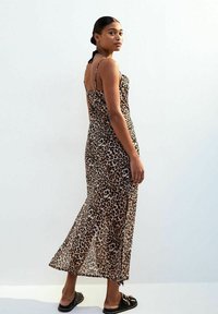 Robe maxi à imprimé léopard avec des fines bretelles, une fente sur le côté et une silhouette fluide. Fabriquée en tissu léger et transparent. Sandales plates présentées.