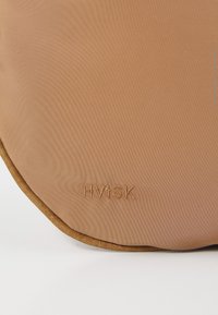 Brun tygväska med en slät textur, rund form och guldbroderi med varumärket "HVISK" nära den nedre kanten.