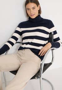 Pull à col roulé à rayures bleu marine et crème avec une texture côtelée, porté avec un pantalon beige, assis sur une chaise moderne noire et chromée.