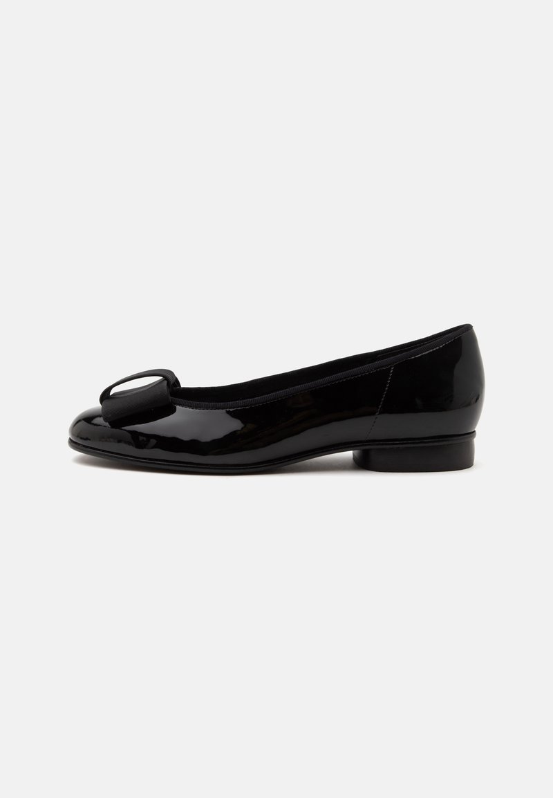 Chaussure plate en cuir verni noir avec un petit talon et un grand nœud décoratif sur l’avant arrondi, sur un fond clair uni.