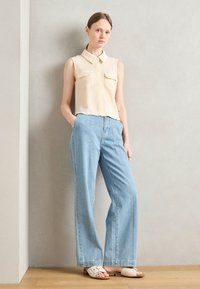 Blusa senza maniche color crema con texture e due tasche sul petto, abbinata a jeans a gamba larga blu chiaro a vita alta e sandali a scivolo color crema.