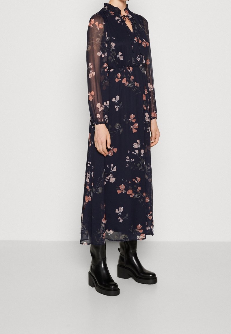 Robe maxi florale bleu marine avec de longues manches transparentes, un col volanté, et des motifs de fleurs roses et blanches, associée à des bottes à talons épais noires.