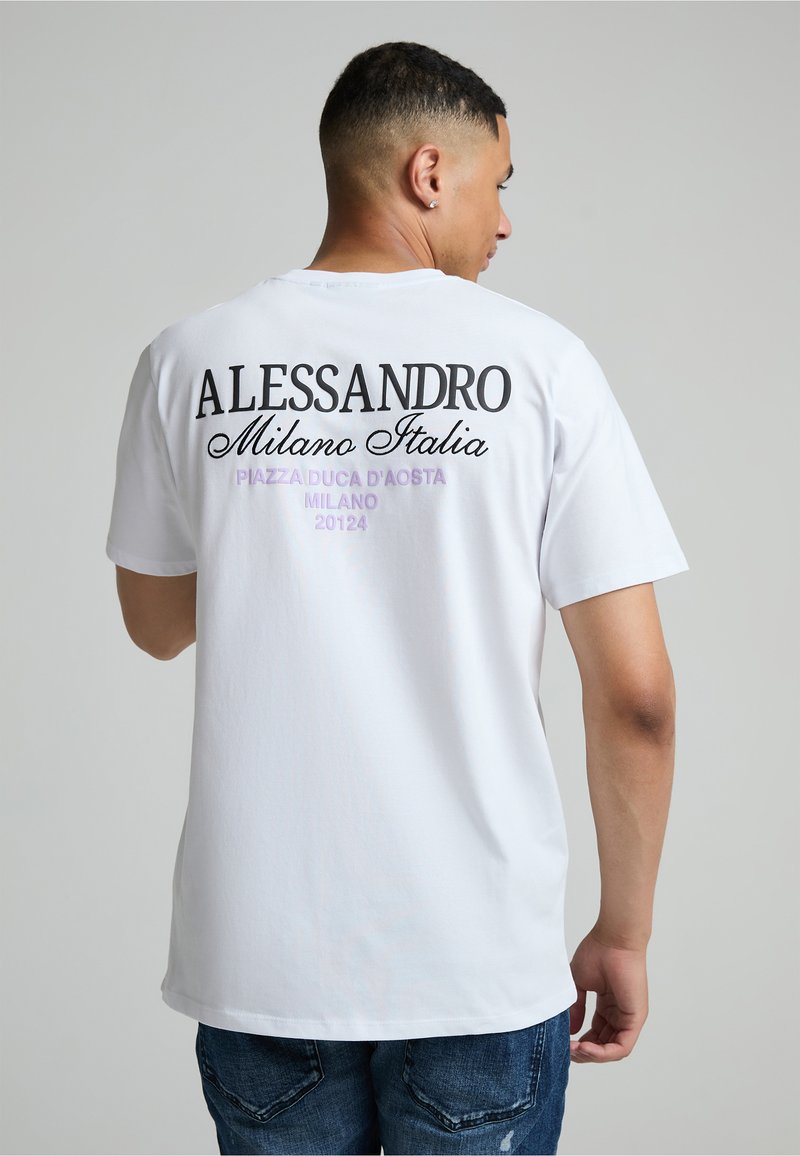 Άνδρας που φοράει λευκό μπλουζάκι με "Alessandro Milano Italia" και ιταλική διεύθυνση τυπωμένη στο πίσω μέρος, σε συνδυασμό με μπλε τζιν.