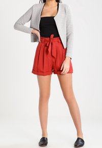 Šedá zkrácená bunda s texturovanou tkaninou, černý top bez rukávů, červené šortky s vysokým pasem a zavazováním, a černé slip-on boty.