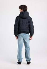 Zwarte gewatteerde jas met capuchon, met een doorgestikt ontwerp, gecombineerd met lichtblauwe spijkerbroek en zwarte sneakers. Achteraanzicht toont geen zichtbare logo's.