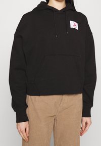 Sudadera corta negra con un bolsillo frontal, capucha con cordón y un parche de logo rojo en el pecho. Hecha de una tela suave con puños acanalados.