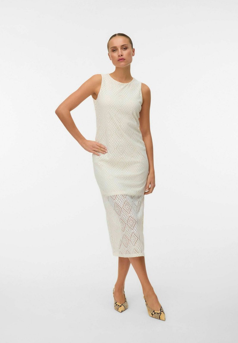Femme en robe midi blanche en dentelle sans manches et escarpins pointus en peau de serpent, debout avec une main sur la hanche sur un fond uni.