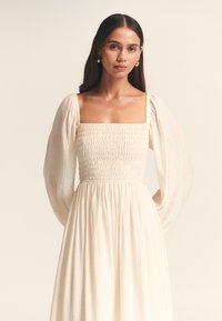 Weißes langes Kleid mit quadratischem Ausschnitt, gerafftem Oberteil und langen, puffigen Ärmeln, gefertigt aus leichtem Stoff mit weicher Textur.