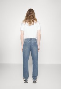 Jeans in denim azzurro chiaro con una vestibilità comoda, cinque tasche e vita alta, abbinati a una t-shirt bianca. Lo sfondo è semplice e chiaro.