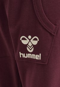 Burgunderfarbener Stoff mit gerippter Textur zeigt ein weißes Logo und den Markennamen "hummel" im Taschenbereich. Einfaches, sauberes Design.