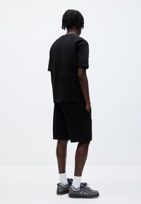 Homme aux cheveux tressés portant un t-shirt noir, un short noir, des chaussettes blanches et des baskets grises, debout les mains dans les poches, de dos.