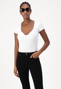 Balta pieguļošā blūze ar īsām piedurknēm, dziļu V veida kakla izgriezumu un teksturētu audumu, kopā ar melniem augstiem džinsiem. Modelis valkā tumšas saulesbrilles.