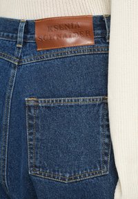 Barna bőrpacsni "KSENIA SCHNAIDER" felirattal a kék farmerekről, amelyeket egy krémszínű kötött felsővel viselnek.